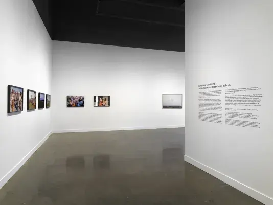 A view of a passageway into a gallery. On the left, six framed photographs showing figures in period dress hang on the wall; on the right is a vinyl introductory exhibition text.