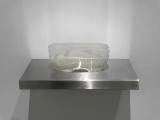 A vaguely boat- or wonton-shaped transparent glass sculpture sits on a metal wall shelf. The sculpture contains an inner space or shape that resembles an abstracted figure, with a rounded head and ovular body.