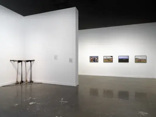 In a corner on the left, a droopy sculpture consists of paired ovular black and brown masses hanging from stick wedged horizontally between the walls. On the right, on a wall further back, framed photographs depict a procession of people carrying flags and weapons. In the center, a sheet of paper with text hangs on the wall.