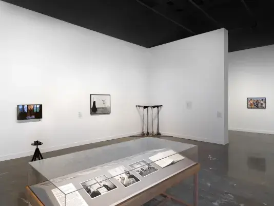 In the foreground, grayscale photographs and an open book occupy a wooden vitrine. The photographs show people in interior spaces, like a man sitting in a kitchen near a stovepipe and a child in a bath near his mother. Other artworks fill the background, including a droopy sculpture, a video monitor depicting a lecture, and a photograph of a procession.
