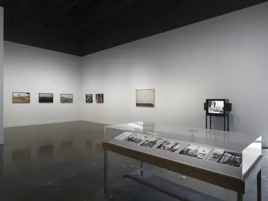 In the foreground on the right, a freestanding wooden vitrine is filled with grayscale photographs: these include a figure sprawled on a chair and a man sitting limply on a porch. Other artworks fill the background, including a boxy monitor depicting a seated woman and a framed photograph of printed pages of text hanging on a wall.