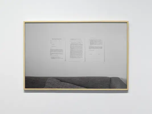 A photograph framed in light wood hangs on the wall. The photograph shows three printed pages hanging, in turn, on a white wall above a couch, only a fragment of which is visible. An unsigned contract is printed on the pages. At the top of the left page, a title is printed in cursive: “Deed of Ownership and Conditions of Existence.”