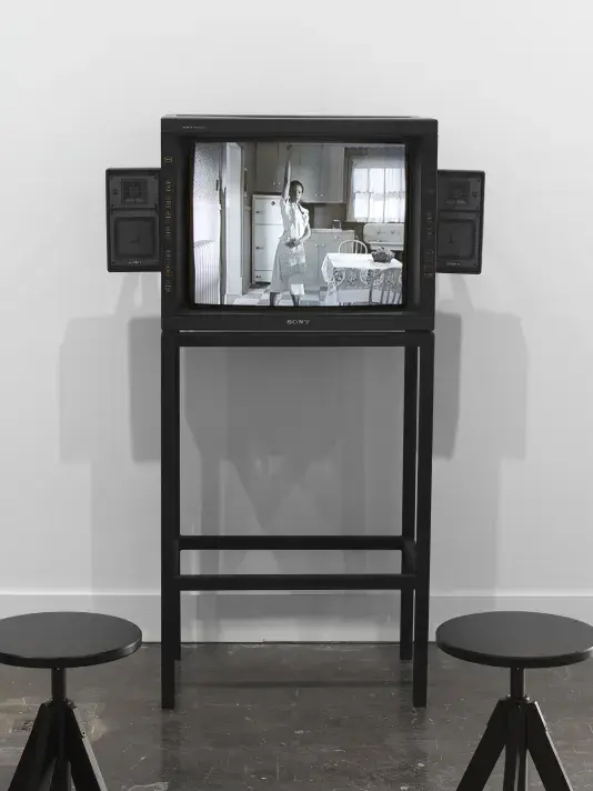 A boxy video monitor with built-in speakers sits on a streamlined metal stand with two stools in front of it. Onscreen, the grayscale image shows a woman wearing white raising her arm straight above her head in a kitchen.
