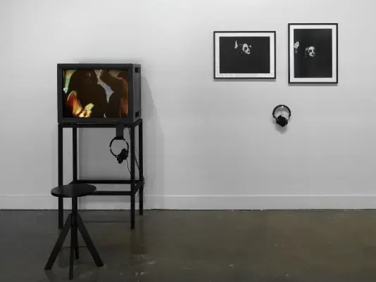 A multimedia installation consists of two parts. On the left, a boxy monitor shows the blurry heads and torsos of dancing figures. On the right, two framed photographs show a woman, her face painted white, wearing sunglasses; little other than her face and right hand are visible. A pair of headphones hangs under the photographs.