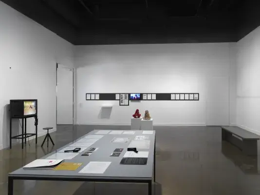 In the foreground, a rectangular table encases documents and drawings—an orange envelope, printed letters, small photographs—under a sheet of clear glass. Handheld magnifiers and a bound volume sit on top of the glass. Other artworks occupy the background, including a boxy monitor showing a dance rehearsal and printed emails hanging in a row on a wall.