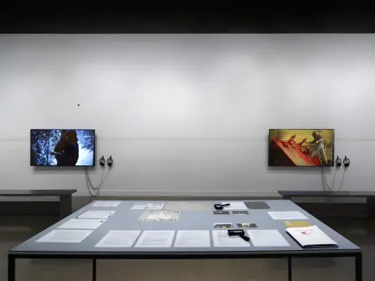 In the foreground, a rectangular table encases documents and drawings—an orange envelope, printed letters, small photographs—under a sheet of clear glass. Handheld magnifiers and a bound volume sit on top of the glass. Two large flatscreen monitors hang on a wall in the background: one shows figures in a snowy landscape; the other shows a factory worker fabricating a red puzzle-like sculpture.