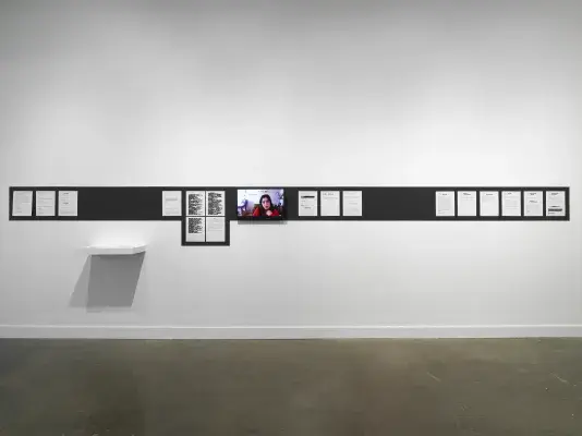 On a white wall, a stripe of black paint outlines printed documents tacked in a row (some of their contents redacted in black rectangles) and a video monitor showing a webcam feed of a woman’s head. A small wall shelf holding two stacks of paper hangs below the stripe, towards the installation’s left side.