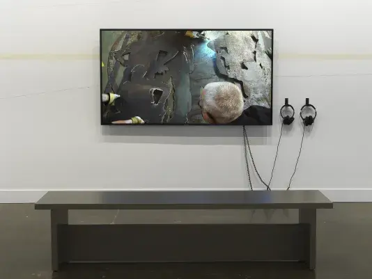 A large flatscreen monitor shows the back of a man’s head and layered silhouettes of posing workers cut from silvery metal. Fluctuating lines—two made in pencil, and one diffuse line made of spray tan—travel across the wall behind the monitor.