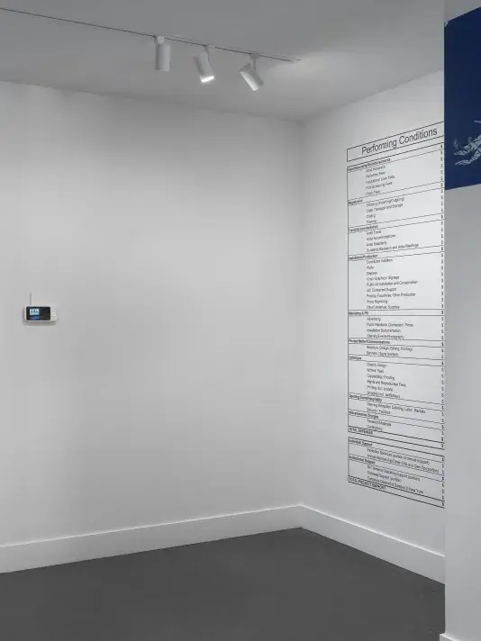 On the left, the screen component of a baby monitor hangs on a wall, dwarfed by the white space around it. On the right, a simple chart labeled “Performing Conditions” is printed on the wall, reaching almost floor to ceiling. The chart is a budget broken up into categories (like Travel/Accomodation and Catalogue) and subcategories (like Writers’ Fees and Shipping).