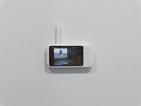 The viewing screen of a baby monitor—with navigation and menu buttons and a white antenna—is attached to a white wall. The feed on the screen shows a person seated on a bench in a dark gallery, watching a projected film.
