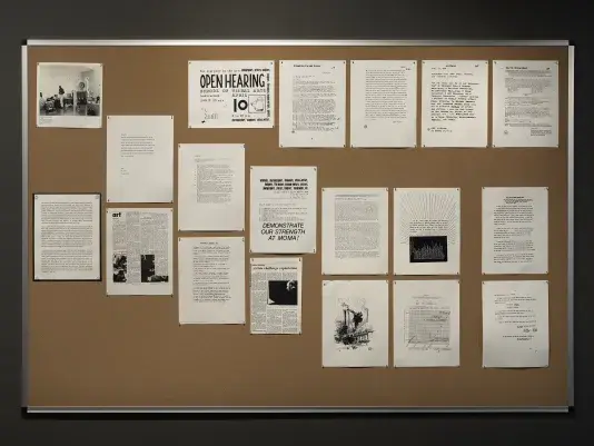 A bulletin board is covered with grayscale documents, mostly letter-sized and covered in text. The largest text is on a flier announcing an “open hearing” at the School of Visual Arts. Another flier enjoins “architects, choreographers … critics, writers, designers” and others to “DEMONSTRATE OUR STRENGTH AT MOMA!”