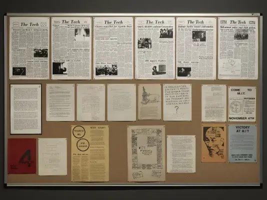 A bulletin board is covered by reproduced documents. In a row at the top, issues of The Tech convey headlines of antiwar protests: “Well-armed police rout I-lab pickets,” reads one. The bulletin board also features copies of posters: one prominently displays the date March 4, while others enjoin readers to come to MIT in November.