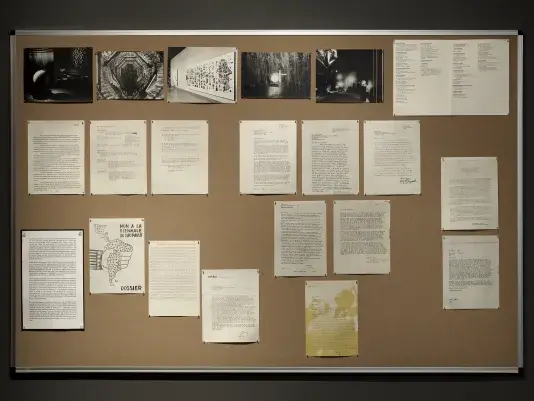 Reproduced documents are arranged on a bulletin board. Most consist of text, but one is a grayscale illustration of the outline of Central and South America, filled in with small crosses. The continent is gripped by a hand with stars and stripes on its cuff. Towards the top of the board, a row of photograph shows an exhibition of technological art: shiny balloons and glowing orbs.