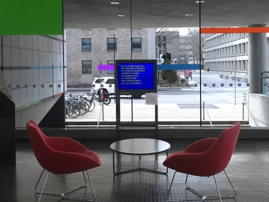 A boxy monitor is positioned against a window, through which a street and the buildings of a campus are visible. The screen shows white text against a bright blue background, reading, “Every dollar spent by the education industry in physical equipment needed to send a message to you is matched by forty dollars spent by you to receive it.”