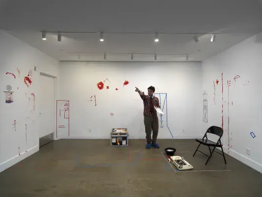 In a gallery, a man wearing a patterned tie holds a printed script in his left hand and points with his right hand. He is surrounded by props and traces of a performance. These include a folding chair, a typewriter, a top hat, and a clown nose on the floor, and obscure geometric figures made of colored tape and more expressive smears of red paint on the wall.