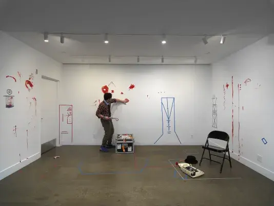 In a gallery, a man wearing a patterned tie holds a printed script in one hand. With the other palm, he smears red paint onto the wall. The walls are already marked by obscure geometric figures—resembling buildings or machines—made of colored tape and other expressive smears and trails of red paint.