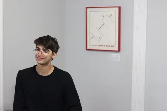 Man wearing a black long sleeve shirt and glasses sits in front of a gray wall with a geometric artwork in a red frame hanging behind.