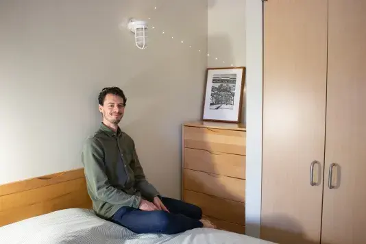 Man in a green button down shirt sits on a bed with a wooden armoire to the right and a black framed black and white photograph leaning on top.
