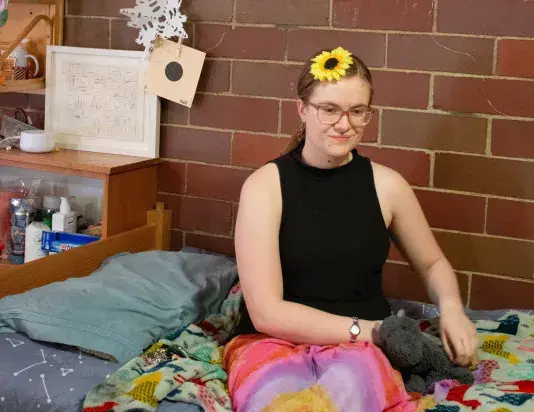 Woman in a black tank top, colorful skirt, glasses and a sunflower in her hair sits on a bed petting a dragon plushy. There is a colorful artwork in a wooden frame to the left of the woman against the brick wall behind.