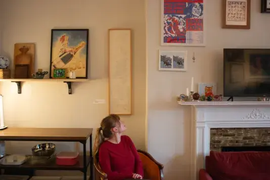 Woman in a red long sleeve top sits in a chair with her hand on her chin in thought. A large rectangular vertical artwork in a wooden frame hangs on the wall behind amongst many other works of art hung in gallery style.