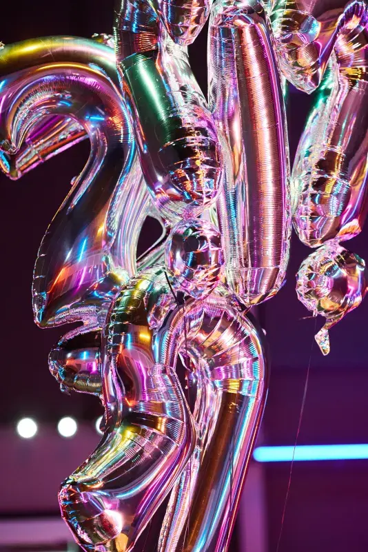 A cluster of shiny, iridescent silver balloons shaped like letters and punctuation marks, including two question marks, reflects pink, purple, and blue light against a dark, blurred background.
