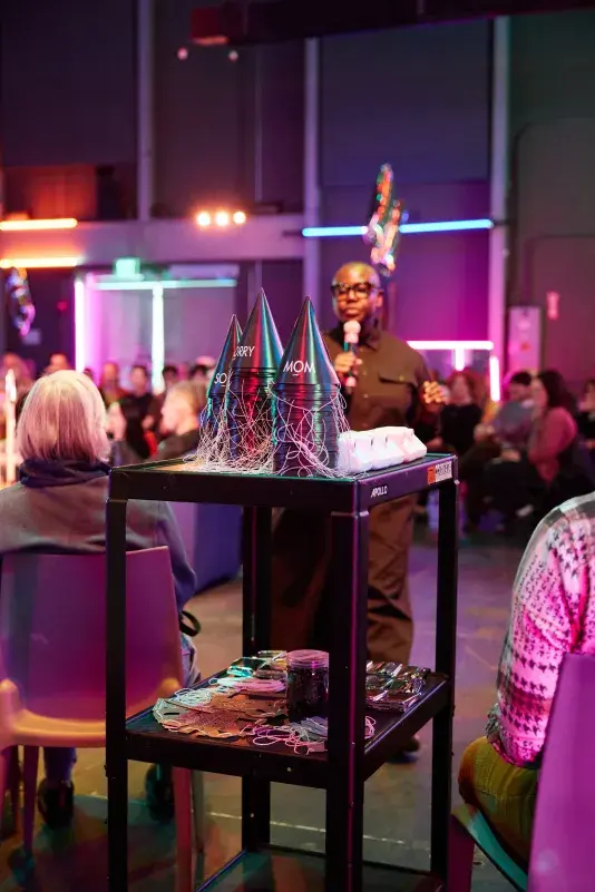 A person in dark clothing and glasses speaks into a microphone, standing behind a black tiered cart. On the cart's top shelf are three stacks of party hats that read "SORRY" and "MOM." An audience is seated in the colorful, neon-lit background.