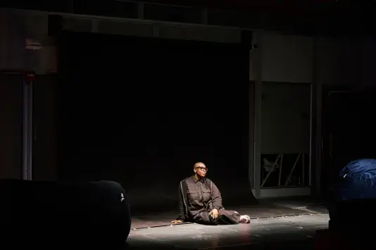 A person wearing black clothing and black glasses sits on a dark stage floor, looking to her left. She sits on a large black backdrop that extends from the wall to the floor, illuminated in a pool of light.
