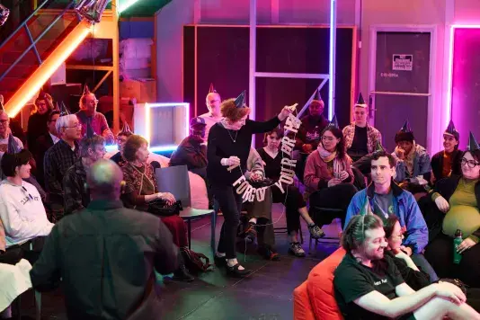 A group of people, many wearing dark party hats, are gathered in a room lit with pink and blue neon lights. A woman with red hair stands up from her seat holding a glittery silver banner of strung letters reading 'FORGOT YOUR BIRTHDAY.’