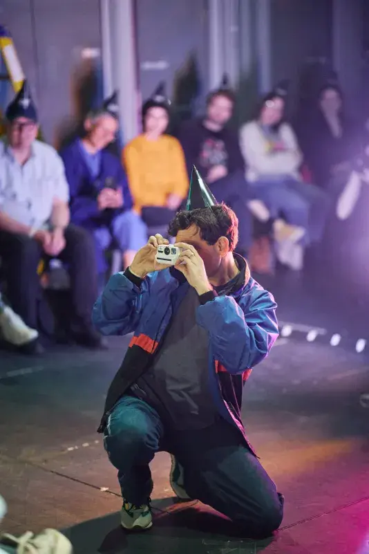A man wearing a black party hat and a blue and black jacket kneels, holding a white instant camera to his face. Several people in party hats are seated in the blurred background.