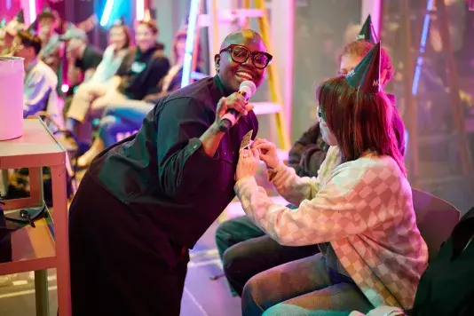 A person with short hair and dark glasses smiles while holding a microphone and leaning towards another person. The second person, wearing a party hat and a checkered sweater, pins a dollar bill to the performer’s shirt. Blurred figures and neon lights are visible in the background.