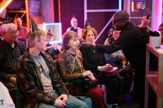 A person holding a microphone stands and feeds a spoonful of food to a seated older woman wearing a patterned cardigan. Several other people in black party hats are seated around them in an indoor setting with neon lights.