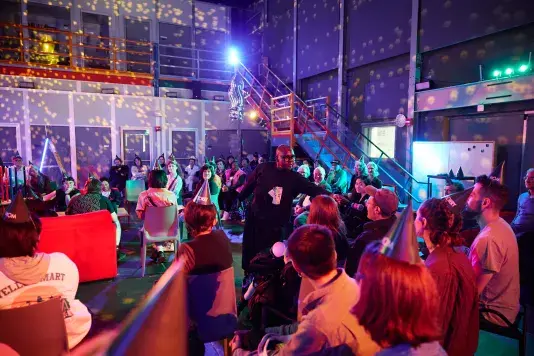 A person in a black outfit and glasses stands in the center of a performance space and holds a microphone to a seated man’s mouth. A few dollar bills are pinned to her chest. Many audience members wear pointed party hats, and the room is illuminated with multicolored lights and disco ball patterns.