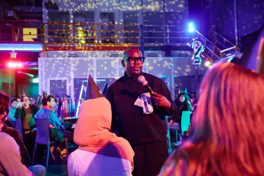 A person with dark-rimmed glasses and a black collared shirt speaks into a microphone. A few dollar bills are pinned to her chest. The room is illuminated by colored lighting and a disco ball, and audience members wearing party hats are visible in the background.