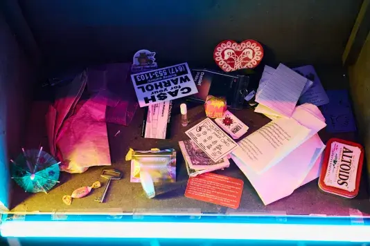 A collection of small items, including a green drink umbrella, a key, a dollar bill, a "CA$H for your WARHOL" sticker, crumpled pieces of paper, and an Altoids tin, are scattered on a cardboard surface