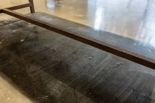 A dark wooden beam, the bottom of a freestanding vitrine, extends diagonally across a floor. Light striations, specks of dirt, and numerous dust bunnies sit on the floor underneath the vitrine.