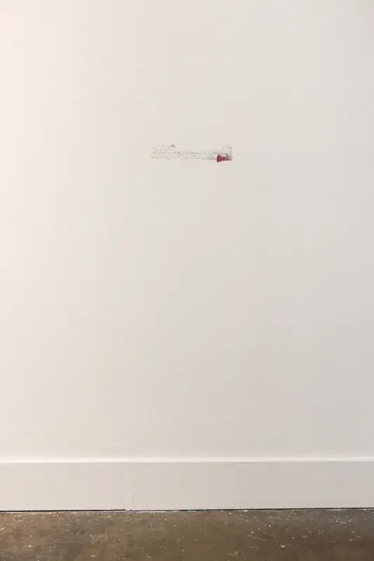 A white wall features a small, horizontal gray residue mark with a magenta paint splash on its right end. A white baseboard runs along the bottom, above a dark, mottled floor.