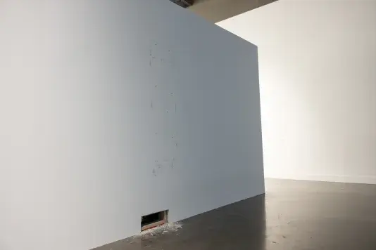 Scuffs and small holes outline an oblong, vertical, rectangular shape on an otherwise bare white wall. Near the floor, a smaller rectangle is cut from the wall entirely, spewing sawdust and scraps of wood onto the floor nearby. A second wall is visible behind the freestanding one.