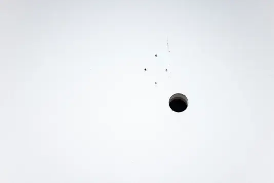 A white wall is punctured by a dark circular hole. Above the hole, four much smaller holes form a diamond shape.