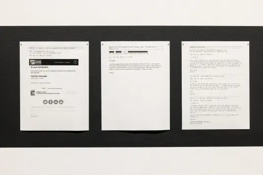 Three printed emails are displayed side-by-side on a white wall, showing correspondence related to a money transfer to someone named Camille; a confused inquiry from a granting agency about an employee’s email signature; and the gallery director’s burnout. The emails are dated April and May 2021.