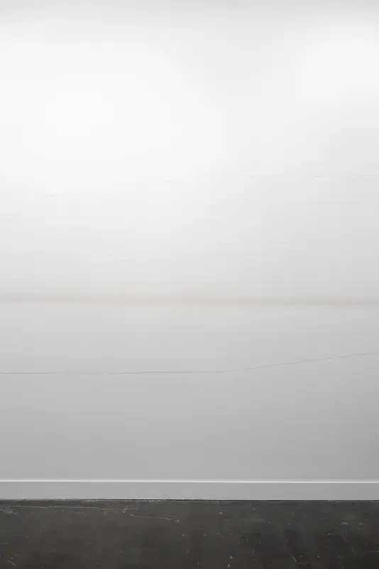 A white wall features three faint, thin horizontal lines that gradually crest upwards and downwards, as well as a more diffuse light brown or gray horizontal band across the middle. The floor in the foreground is dark gray.