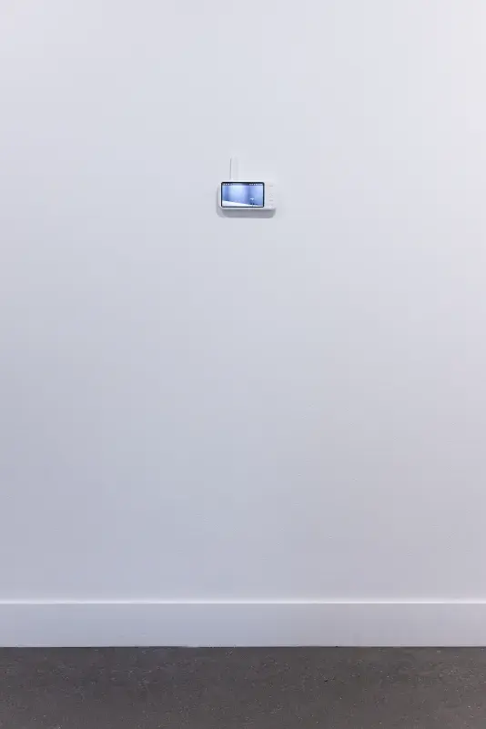 The viewing screen of a baby monitor—with navigation and menu buttons and a white antenna—is attached to a white wall. It is dwarfed by an expanse of white wall and a sliver of gray floor. The feed on the screen shows a single metal stool.