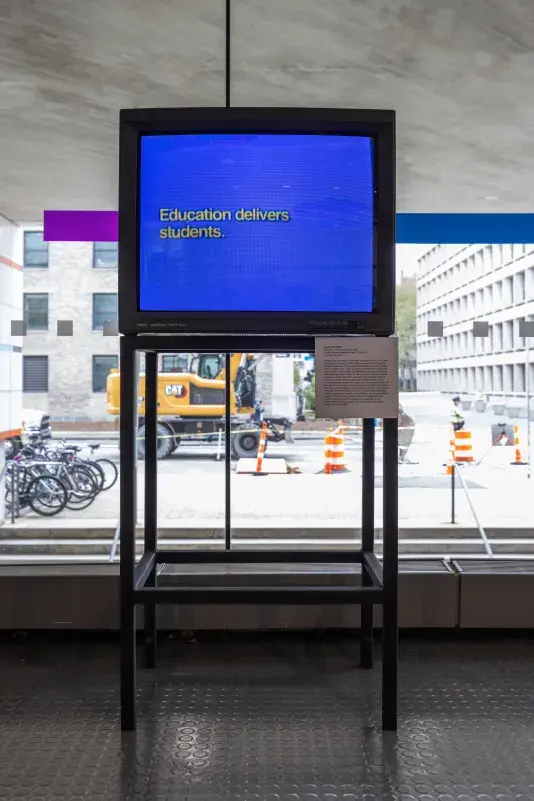 A boxy monitor is positioned against a window, through which a street, construction equipment, and the buildings of a campus are visible. The screen shows white text against a bright blue background, reading, “Education delivers students.”