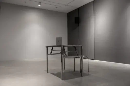 Two pieces of spare, angular, metal furniture occupy an otherwise empty space with gray walls and a concrete floor: a small table and a stool. An open Mac laptop sits on top of the table.