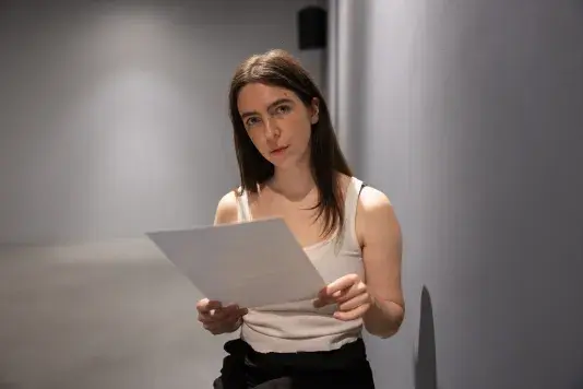 A woman wearing a white tank top and black pants makes eye contact with the camera and holds some papers in her hands. She appears to occupy a bare space with gray floor and walls.