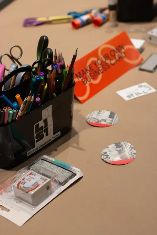 A table covered in brown craft paper with art supplies scattered on top and a red paper sign with black text that reads "MAKE YOUR OWN BUSINESS CARD".