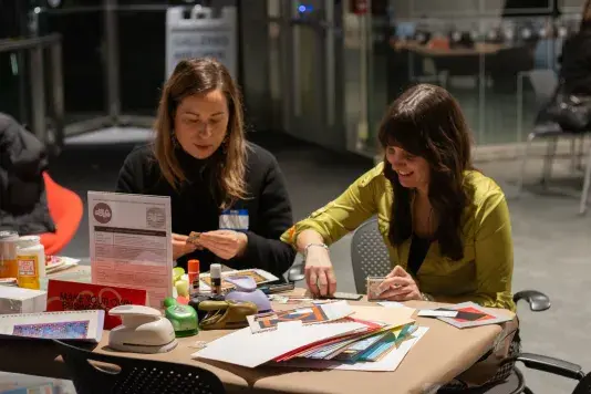 Two people sitting at a table covered in brown craft paper and art supplies working on various crafts.