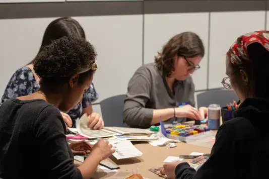 Four people sitting around a table covered in brown craft paper and art supplies working on various crafts.