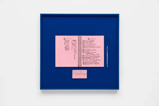 Two pink notecards with handwriting in black pen on top of a dark blue background inside of a black frame that hangs on a white wall.
