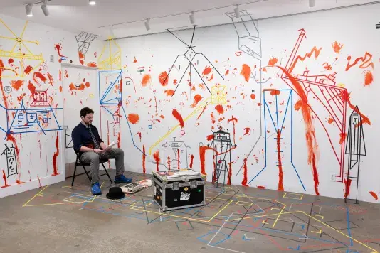A man sits on a sturdy chest or box and looks down at the packet of paper in his hands. The white walls behind him are marked with dense lines of colored tape and splotches and smears of red paint. Other objects, like a hat and a typewriter, are arranged at his feet.