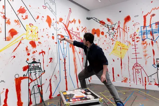 A man wearing a tie stands in a lunge position, one hand pointing at a splotch of red paint on a white wall. The rest of the wall is covered with dense lines of colored tape and splotches and smears of paint.